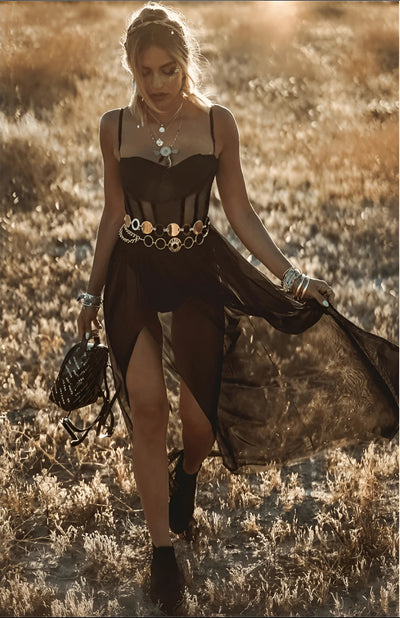 Woman wearing a Bohemian sheer black dress with a silver chunky belt, smiling on a beach with the sky in the background.